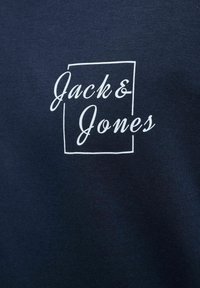 Tamsiai mėlynas medvilninis marškin�ėliai, turintys baltą spaudinį "Jack & Jones" stačiakampio kontūre. Lygus paviršius su klasikinio apvalaus kaklo dizainu.