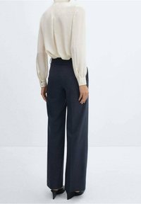 Žena v tmavých širokých kalhotách s vysokým pasem, krémové blůze s dlouhým rukávem zaříznuté do kalhot a černých lodičkách s špičatou špičkou, stojící na jednobarevném bílém pozadí.