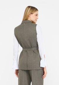Gilet grigio a spina di pesce con un design strutturato, una cintura in vita e un dettaglio con bottone sul retro, abbinato a una camicia bianca a maniche lunghe.