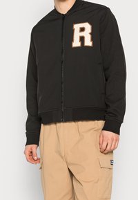 Svart lättvikts bomberjacka med frontdragkedja, ribbade muddar och en applikation av ett "R" på vänster bröst. Har en mjuk tygtextur.
