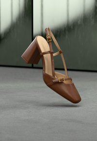 Chaussure à talons hauts en cuir marron avec un bout carré. Elle est dotée de brides autour de la cheville et de boucles dorées, avec une finition brillante et une surface texturée.