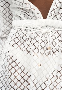 Λευκό δαντελένιο φόρεμα με σχέδιο διαμαντιού και λευκό κορδόνι που δένει φιόγκο στη μέση με μεταλλικά τελειώματα.