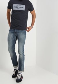 Námorníkově modré tričko s krátkým rukávem s šedým obdélníkovým potiskem, světle modré džíny s ohrnutými nohavicemi a černé tenisky s červenými a bílými akcenty.