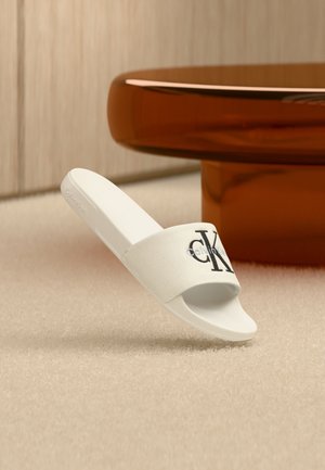 Λευκή σαγιονάρα Calvin Klein με μαύρο λογότυπο "CK" σε υφασμάτινο λουράκι, τοποθετημένη σε μπεζ χαλί κοντά σε στρογγυλό τραπέζι χρώματος κεχριμπαριού.