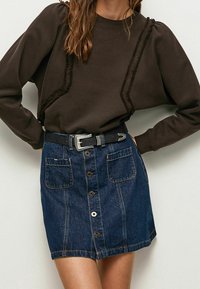 Sudadera marrón con detalles de ribete texturizado combinada con una falda de mezclilla azul claro que presenta un cierre de botones en la parte delantera y dos grandes bolsillos. Cinturón negro incluido.