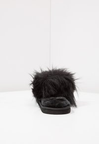 Black fur slipper with a thick, fuzzy upper and soft flat sole. The texture is plush, featuring a high, shaggy exterior and smooth interior.