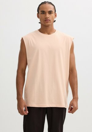 Hombre con cabello trenzado, vistiendo una camiseta sin mangas beige y pantalones negros, de pie frente a un fondo liso.