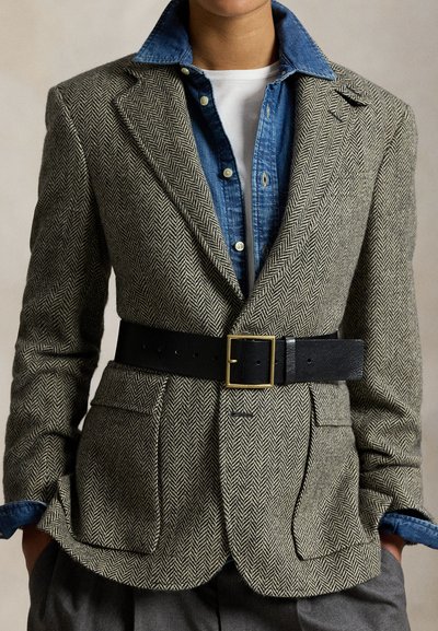 Osoba nosící šedý blazer se vzorem rybí kosti s černým pásem přes modrou džínovou košili a bílým tričkem, ruce v kapse.