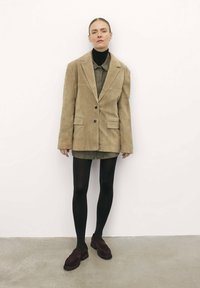 Bežový semišový oversize blejzer s dvoma prednými vreckami, nosený cez olivovo zelenú košeľu na gombíky. Čierne pančuchy a tmavočervené loafers dopĺňajú vzhľad.