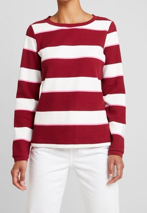 Persoon draagt een trui met lange mouwen en een gestructureerd patroon met brede horizontale rode en witte strepen, gecombineerd met een witte broek.