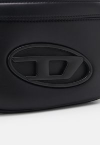 Czarna torba z ekoskóry z teksturowaną powierzchnią i wypukłym owalnym logo w kolorze czarnym. Projekt kładzie nacisk na minimalizm i branding.