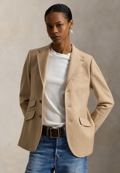 Beige Wollblazer mit Notch-Revers, zwei Fronttaschen und Knopfverschluss, getragen über einem weißen T-Shirt und blauen Jeans.