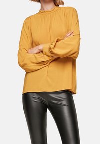 Senapsgul blus med samlad midja, långa ärmar och knappar, i kombination med svarta tajta byxor. Mjukt tyg och avslappnad passform.