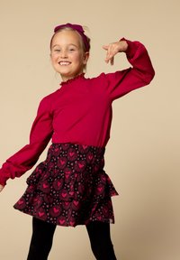 Lachend meisje in een rode lange mouw top, zwarte rok met rode hartpatronen, zwarte leggings en een rode haarband, poserend met opgetrokken armen.