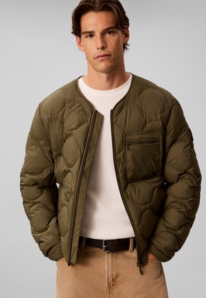 Jeune homme portant une veste matelassée vert olive sur une chemise blanche et un pantalon beige, les mains dans les poches, sur un fond gris uni.