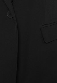 Blazer nero con una texture liscia, un bottone singolo e una tasca angolata alla moda sul lato sinistro. Design semplice ed elegante.