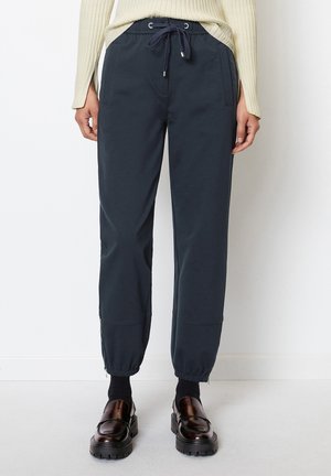 Woman wearing navy drawstring jogger pants with elastic cuffs, black socks, brown loafers, and a cream ribbed long-sleeve top.