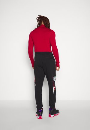 Osoba z dredami stoi przodem do ściany, ubrana w czerwony długi rękaw, czarne spodnie z graficznymi naszywkami oraz czarne trampki z czerwonymi podeszwami.