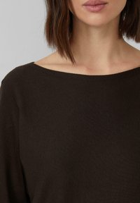 Femme aux cheveux bruns raides portant un pull ample marron foncé, découvrant les épaules et une partie du cou et des lèvres.
