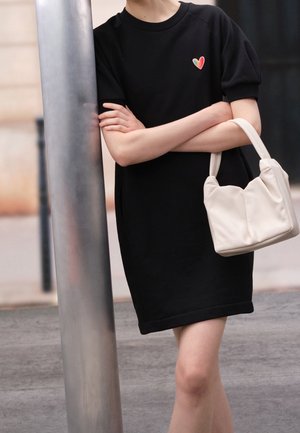 Person wearing a black dress with a small rainbow heart patch, arms crossed, holding a white handbag, standing next to a metallic pole outdoors.