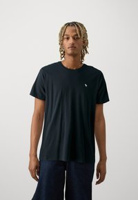 Camiseta de algodón negra con mangas cortas, cuello redondo y pequeño logo blanco en el pecho, combinada con pantalones de mezclilla azules holgados.