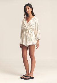 Hvid slå-om-style romper med sorte polkaprikker, lange puffede ærmer, indsnøret talje og en sløjfedetalje. Kombineret med sorte flip-flops.