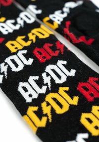 Calzini neri con testo ripetuto "AC/DC" in bianco, giallo e rosso, con un accento a forma di fulmine. Materiale in maglia morbida con un motivo audace.
