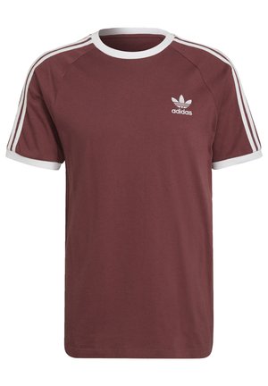 Mężczyzna ubrany w fioletowawy t-shirt Adidas z krótkim rękawem i pasujące spodnie z białymi paskami na ramionach oraz logo Adidas na piersi i udzie.