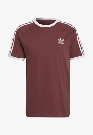Mężczyzna ubrany w fioletowawy t-shirt Adidas z krótkim rękawem i pasujące spodnie z białymi paskami na ramionach oraz logo Adidas na piersi i udzie.