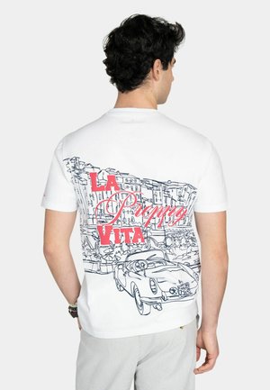 Hombre con cabello rizado y oscuro, llevando una camiseta blanca, mostrando un boceto de edificios y un coche clásico con el texto rojo "LA DOLCE VITA" en la espalda.