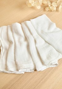 Baltas kokvilnas muslīna audums ar nedaudz raupju virsmu, kārtīgi sakārtots maigos krokās uz gaiša koka virsmas.