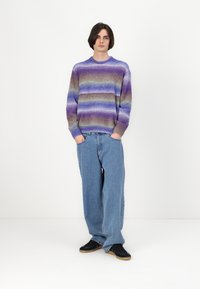 Gebreide trui in paarse gradient strepen, met een ronde hals en geribde mouwen, gecombineerd met lichtblauwe baggy jeans en zwarte sneakers.