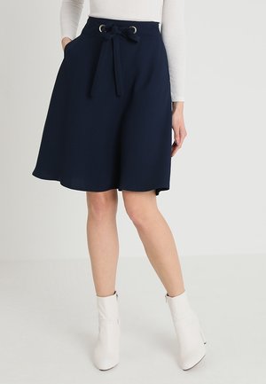 Femme portant une jupe bleu marine longueur genou avec une ceinture nouée et des poches, associée à des bottines blanches et un haut blanc à manches longues.