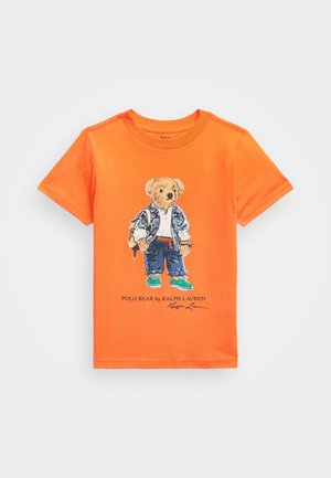 Fel oranje T-shirt met korte mouwen met een beer die een jas, spijkerbroek, sneakers en een rugzak draagt, bedrukt op de voorkant, gesigneerd "Polo Bear by Ralph Lauren."
