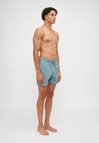 Lyseblå svømmebukser med elastisk linning, snor og glatt tekstur. Modellen står barbeint og viser en sideprofil.