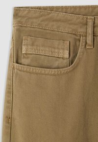 Pantaloni di cotone beige con una tessitura diagonale e texture. Presentano una tasca sul retro e passanti per cintura lungo la vita. Design pulito e semplice.