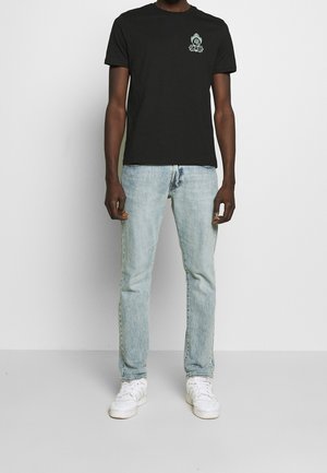 Personne portant un t-shirt noir avec un petit emblème, un jean bleu clair et des baskets blanches, se tenant contre un fond blanc uni.