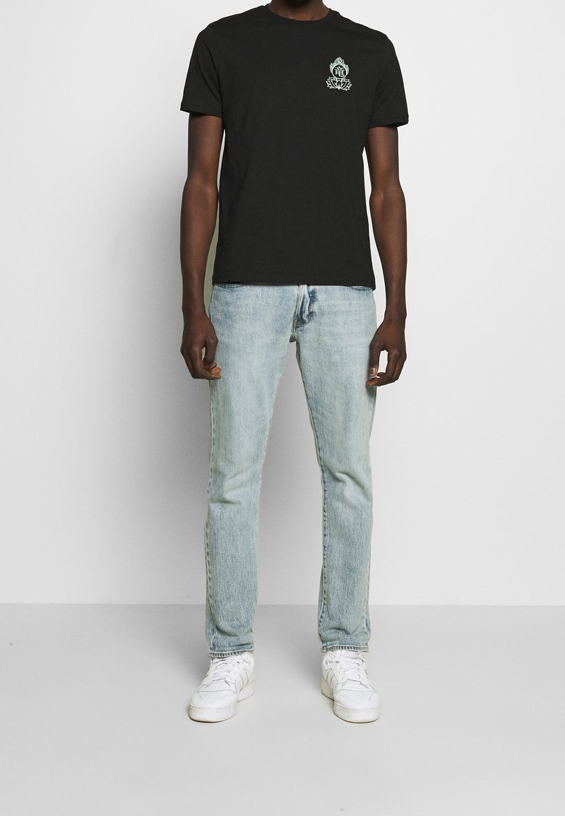 Personne portant un t-shirt noir avec un petit emblème, un jean bleu clair et des baskets blanches, se tenant contre un fond blanc uni.