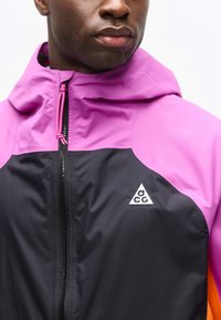 Άνδρας που φοράει μαύρο και ροζ μπουφάν με κουκούλα, με φερμουάρ μπροστά και ένα λευκό τριγωνικό λογότυπο ACG στο στήθος.