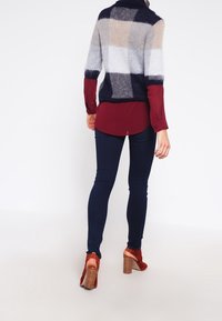 Person som bär en rutmönstrad fluffig tröja, burgundisk blus, mörka smala jeans och bruna högklackade sandaler står mot en vit bakgrund.