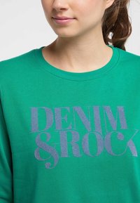 Sudadera verde con cuello redondo y texto impreso "DENIM & ROCK" en gris. La tela parece suave y de peso medio.