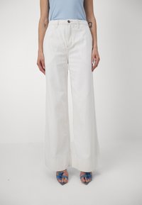 Pantalons larges blancs en denim, avec taille haute, fermeture par bouton à l'avant et lignes épurées. Associés à des sandales bleues.