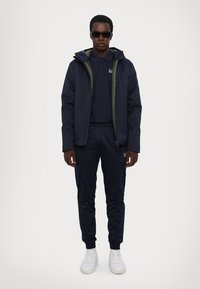 Giacca puffer blu navy con foderatura verde, indossata sopra un polo blu navy e pantaloni jogger abbinati, abbinata a sneaker bianche e occhiali da sole neri.