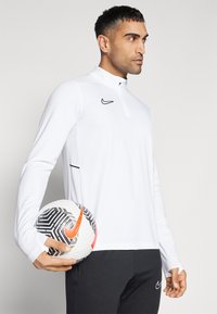 Weißes Nike Langarm-Sportoberteil mit einem halben Reißverschluss, schwarzem Logo und seitlichen Mesh-Akzenten. Das Model hält einen schwarz-weiß gemusterten Fußball.