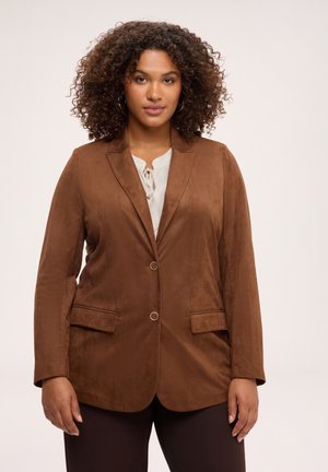 Donna con capelli ricci che indossa un blazer marrone sopra una camicetta chiara, in piedi davanti a uno sfondo neutro semplice.