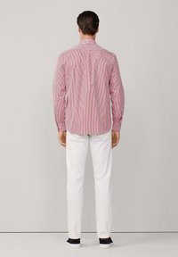 Un homme portant une chemise à rayures rouges et blanches à manches longues, un pantalon blanc et des chaussures noires, se tenant face à un fond gris clair uniforme.