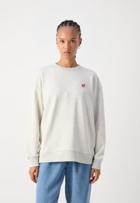 Ανοιχτό γκρι φούτερ σε μαλακό ύφασμα, με στρογγυλή λαιμόκοψη, χαμηλούς ώμους και ένα μικρό κόκκινο λογότυπο στο αριστερό στήθος.