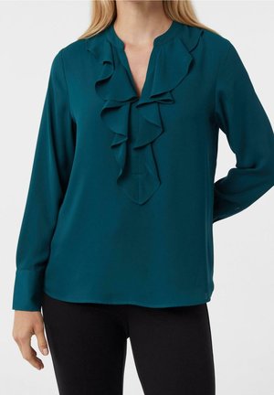 Kvinde iført teal langærmet bluse med lagdelt flæsedetalje ved V-hals, parret med sorte bukser mod en ensfarvet baggrund.