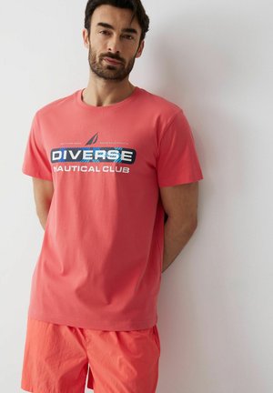 Mężczyzna o ciemnych włosach i brodzie, ubrany w koralowo-różową koszulkę "Diverse Nautical Club" i pasujące szorty, stoi przy białej ścianie.