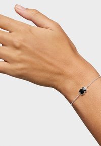 Pulsera con charm en forma de trébol de cuatro hojas en una muñeca, que presenta una delicada cadena de plata y una textura suave. Diseño simple con un acabado pulido.